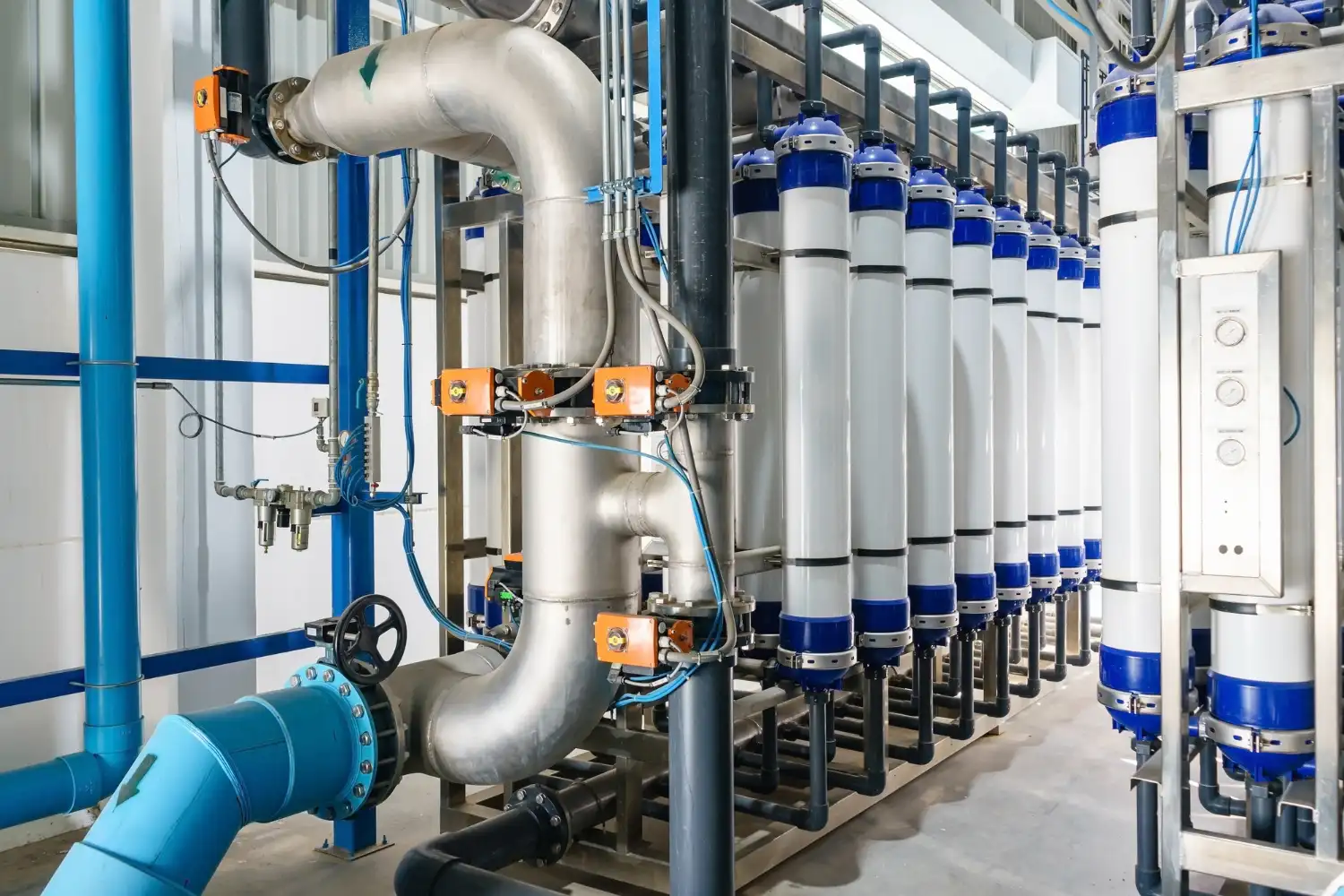 Sala industrial com tubulações, válvulas e conjunto de cilindros brancos com tampas azuis, equipado com dosador de cloro gás para tratamento e dosagem automatizada.