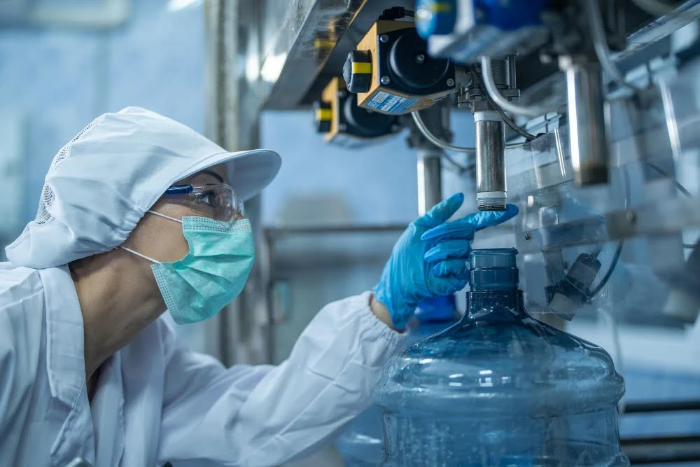 Profissionais em laboratório com máscaras e luvas operam equipamento industrial de tratamento de água farmacêutica, preparando e controlando frascos em ambiente estéril com tubulações e sensores.