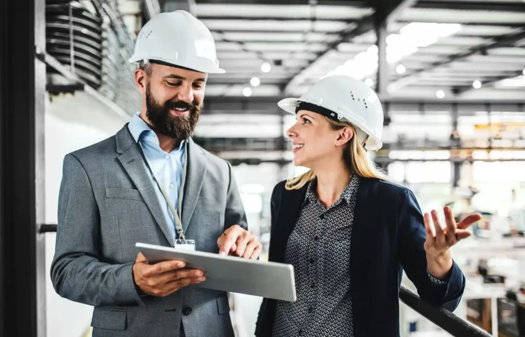 Engenheiro e supervisora usando capacetes brancos em uma fábrica, analisando um tablet e conversando no ambiente industrial, com foco em eficiência operacional e custo total de propriedade