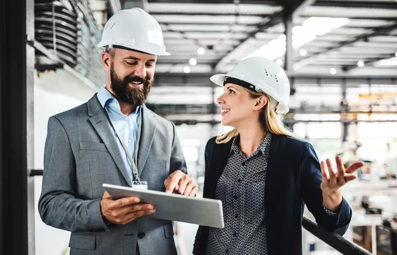 Engenheiro e supervisora usando capacetes brancos em uma fábrica, analisando um tablet e conversando no ambiente industrial, com foco em eficiência operacional e custo total de propriedade