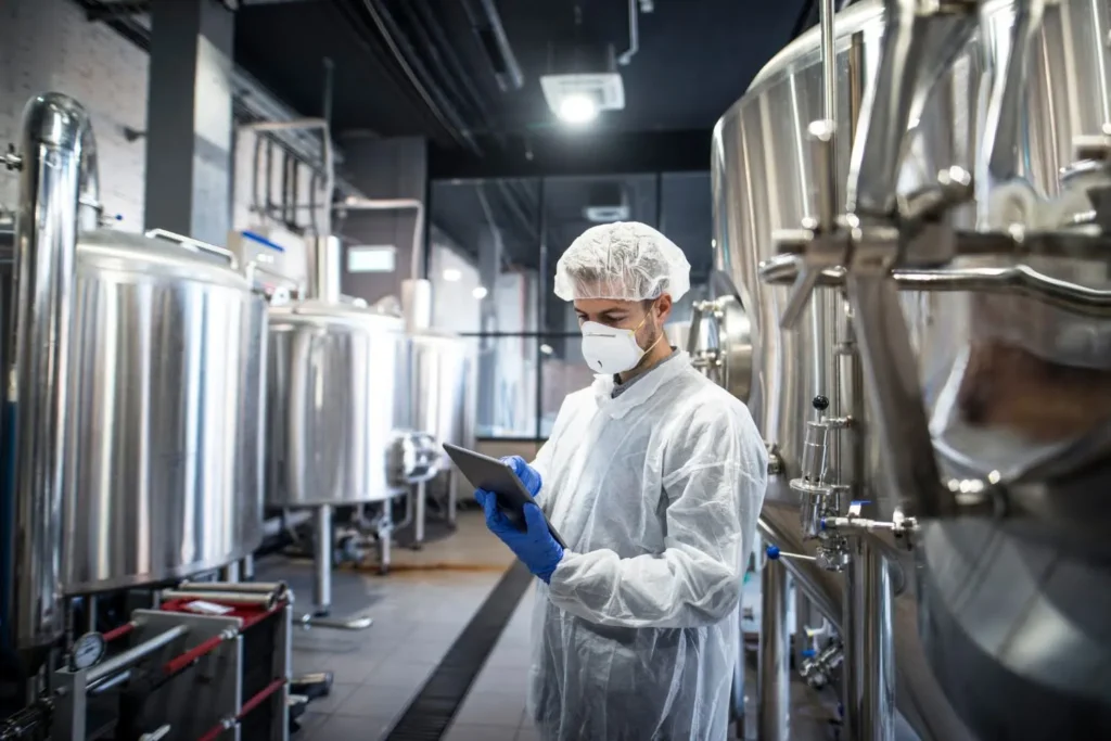 Profissional em laboratório de produção industrial, usando jaleco e máscara, inspeciona com um tablet equipamentos de aço inox em uma fábrica, com foco em monitoramento da qualidade da água